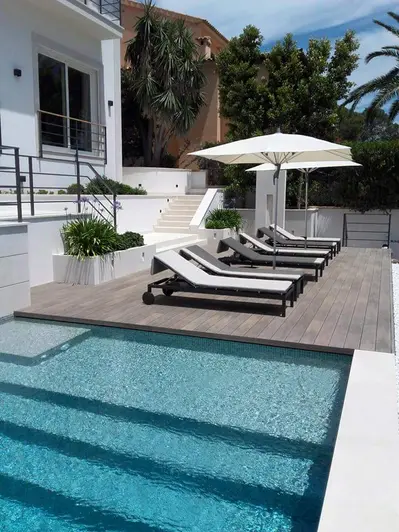 Poolside view with 6 sunloungers sit on traditional designboard composite decking, white clad house and palm trees in the distance.