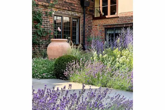 A pithoi rises above dense floriferous planting edging Tumbled Black Indian sandstone paving outside period brick and tile-hung house.