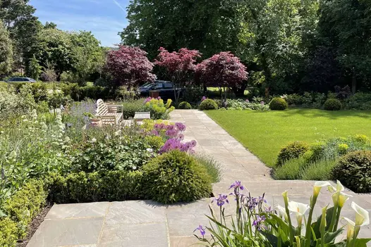 Tumbled Black Sandstone paving runs between flowerbeds and well manicured lawn, bench and table to the left with trees surrounding.