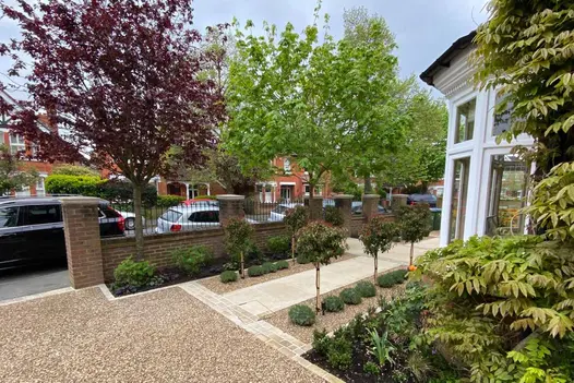 Path to the front door featuring a cross-shaped design bordered by Tumbled Mint Sandstone Setts, framed by pebbles and bushes.