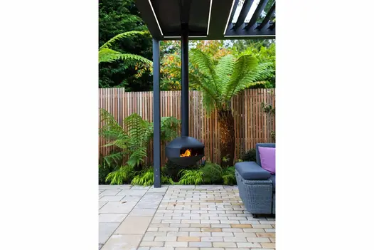 Paving slabs meet tumbled mint sandstone setts laid running bond, under corner of louvred metal pergola with hanging woodburner.