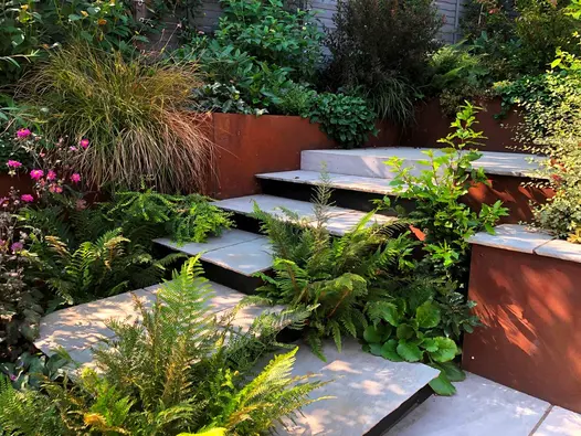 Steps of tumbled Kandla grey garden paving slabs turn right angle and rise to paved area surrounded by lush planting.