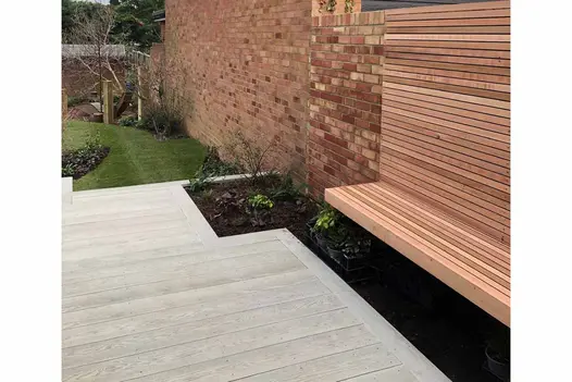 Floating bench sits on brick wall with flowerbeds underneath, smoked oak millboard decking leads down to lawn area.