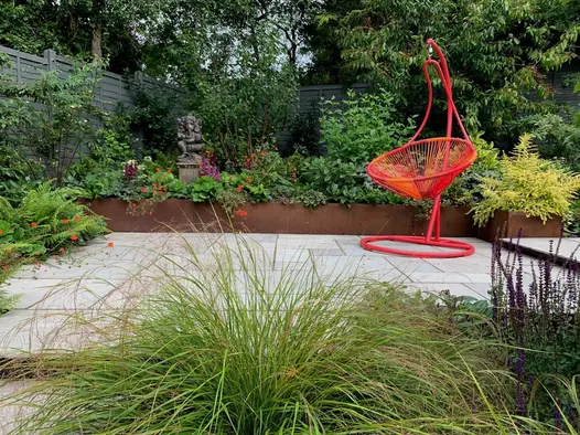Hanging chair on rectangular patio of Kandla Grey tumbled Indian sandstone slabs, with raised beds of Corten steel lushly planted.