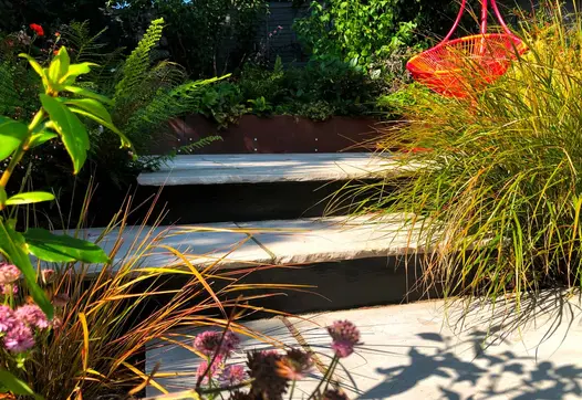 2 Kandla Grey tumbled sandstone step with rock-hewn edges rise between planting toward orange hanging chair.