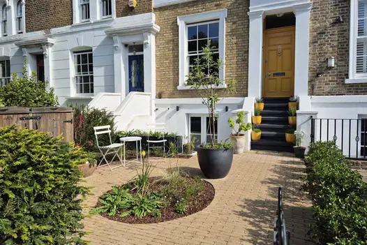 Patio of Westminster Clay Pavers has circle cut out for flowerbed, leading up to front door of house, with white metal furniture,