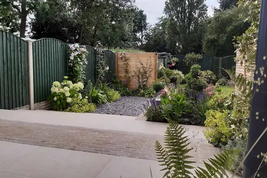 Westminster Clay Pavers indent porcelain patio with flowerbeds against green fencing, steps lead down to pebbled area.