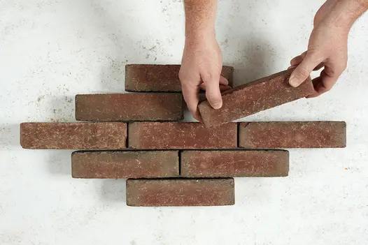 Against plain white background, 2 hands insert a 9th Winton clay paver to the 2nd row of a lozenge-shaped arrangement of bricks.